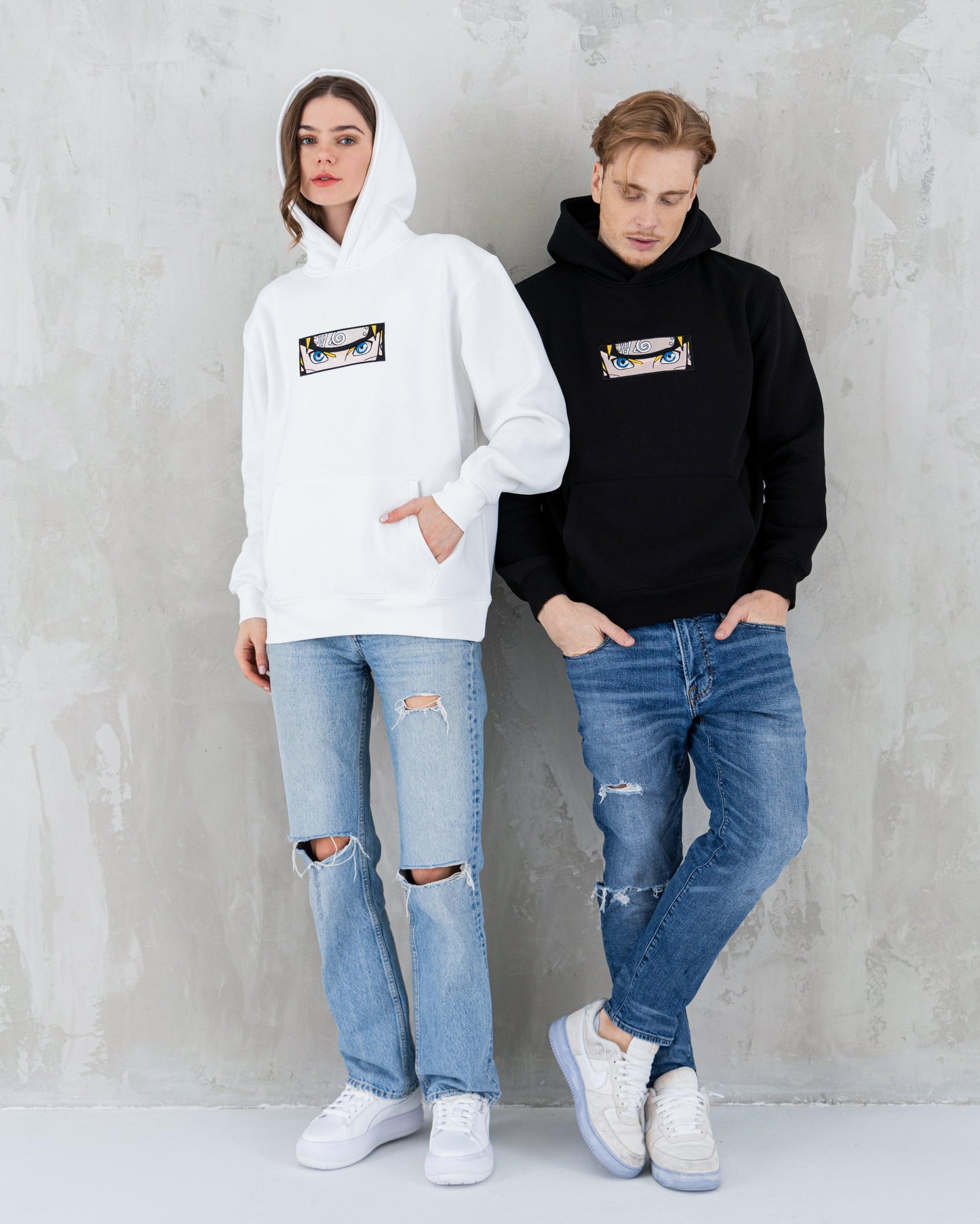 Man and woman wearing anime-themed hoodies with eye graphic, paired with ripped jeans and sneakers.