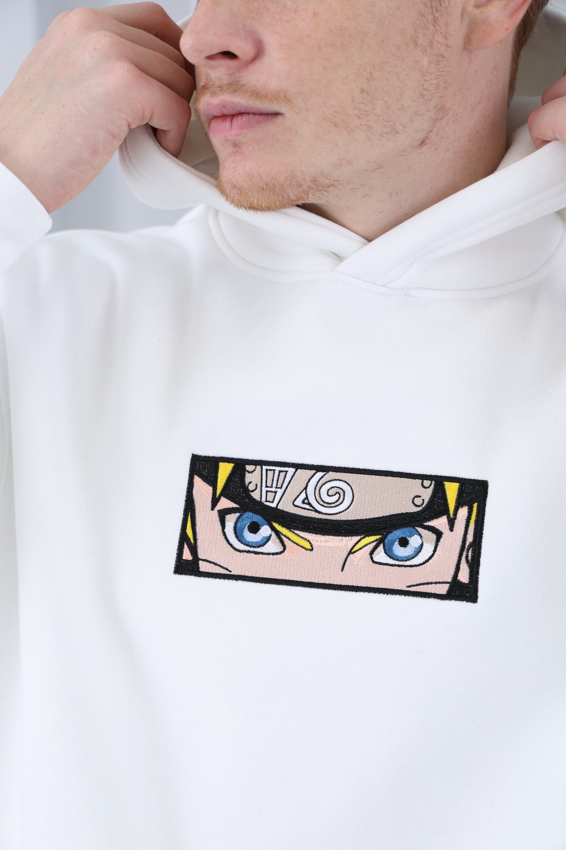 A close-up of a white naruto uzumaki hoodie featuring a detailed anime-style graphic of Naruto Uzumaki's intense blue eyes and forehead protector with the Hidden Leaf Village symbol. The person wearing the hoodie is holding the edge of the hood, showing part of their face with light stubble. The hoodie’s fabric appears soft and smooth.