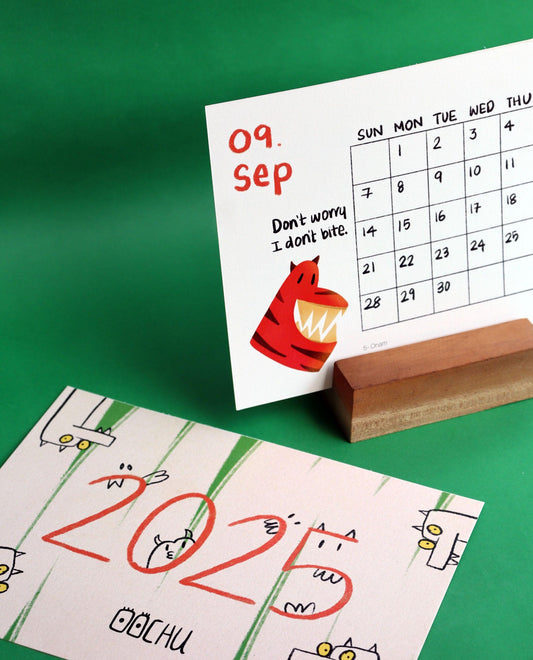 A colorful 2025 calendar design by Oochu. The September page features a cute red tiger illustration with the text 'Don't worry, I don't bite,' next to a simple calendar layout. The foreground showcases a 2025 cover design with playful cartoon-like creatures on a striped background. Both designs are displayed against a vibrant green background, with the calendar card resting on a wooden holder.