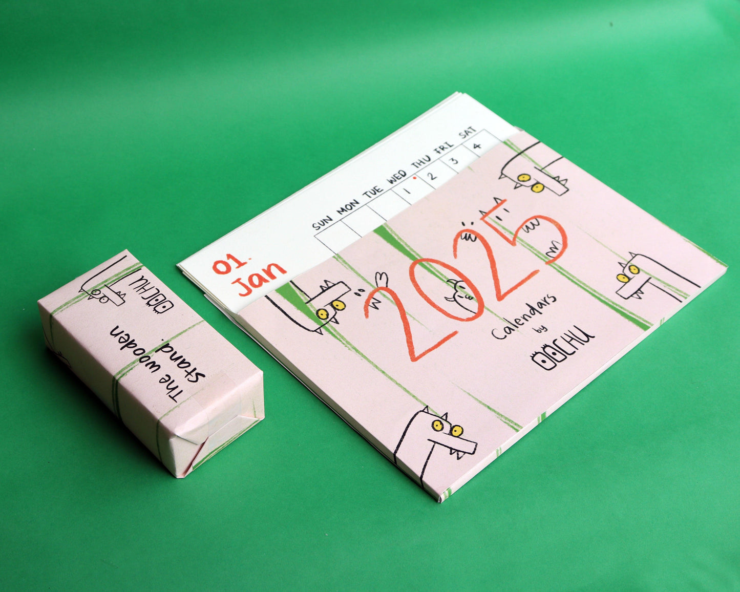 A 2025 calendar set by Oochu, featuring a playful design. The calendar cover has whimsical illustrations of quirky cartoon creatures on a soft pink background with green stripes, and the year '2025' is written in bold red text. A smaller box labeled 'The Wooden Stand' with matching artwork is placed next to the calendar. Both items are displayed against a vibrant green background, highlighting their creative and fun design.